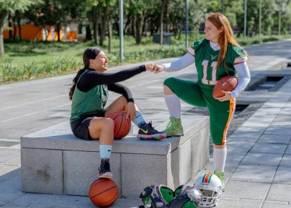 Two female athletes share a fist bump while sitting outdoors, holding sports equipment in Mexico City.